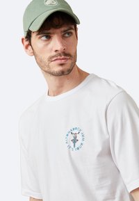 Camiseta blanca de algodón con un logotipo gráfico circular azul con texto, acompañada de una gorra verde. El diseño es simple y casual.