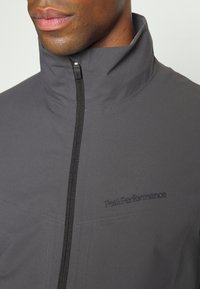 Grijze waterdichte jas met een hoge kraag, een volledige ritssluiting aan de voorkant en het geborduurde "PeakPerformance"-logo in een strak, modern ontwerp.