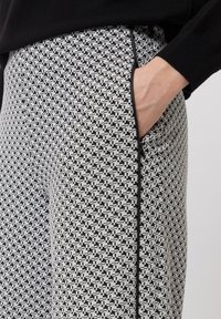 Hand in de zak van zwart-wit gestructureerde broek met zwarte piping, gedragen met een zwart langemouwtop tegen een neutrale achtergrond.