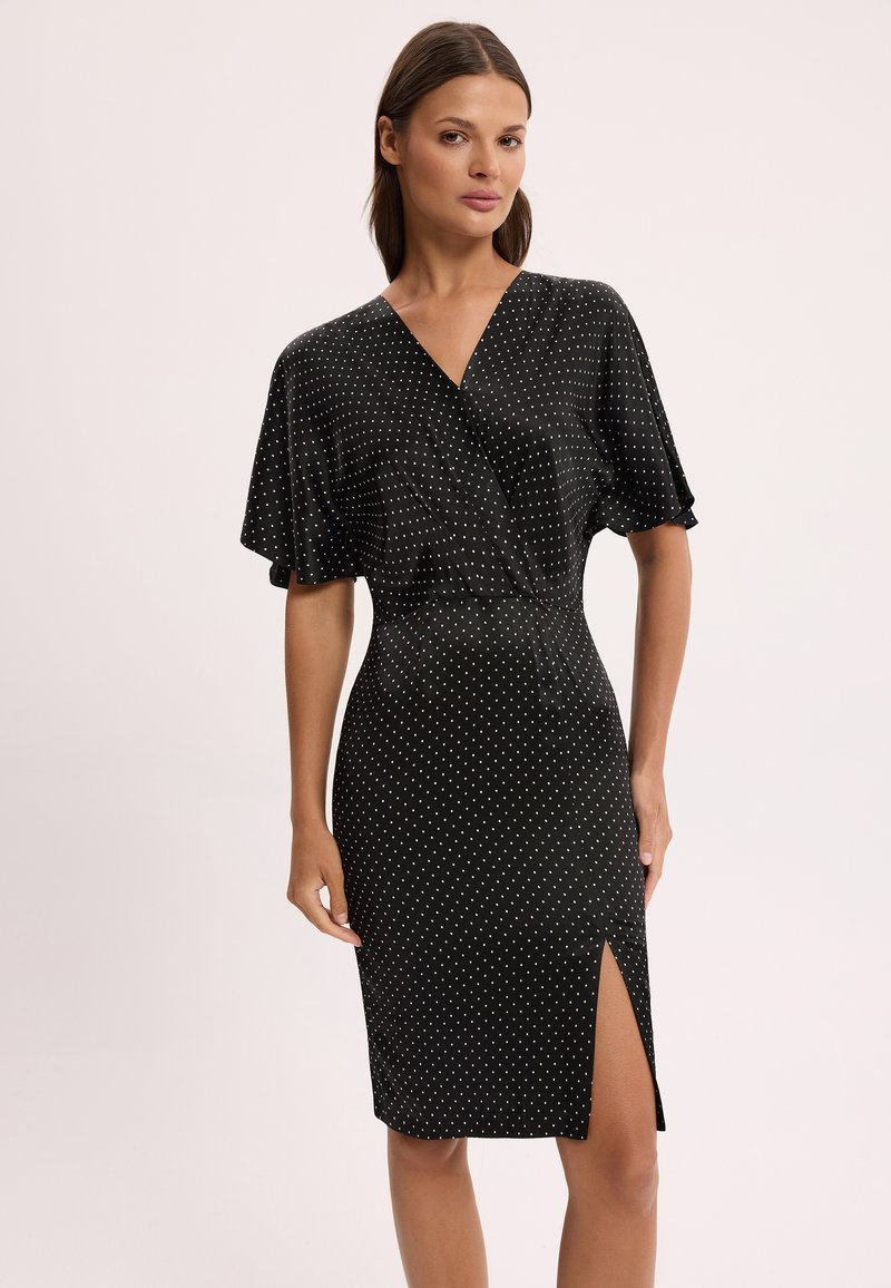 Kobieta ubrana w czarną sukienkę do kolan w białe grochy, z dekoltem w serek, krótkimi szerokimi rękawami i rozcięciem na przodzie na wysokości uda, stojąca na neutralnym tle.