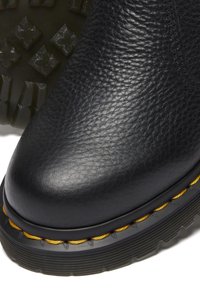 Bota de cuero negra con superficie texturizada, punta redondeada, suela de goma acentuada con costuras amarillas y un diseño de banda de rodadura resistente.