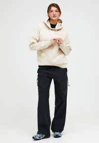 Felpa beige con cappuccio e cordino, con tasca frontale, abbinata a pantaloni cargo neri con tasche con zip e vestibilità ampia. Sneakers con dettagli grigi.