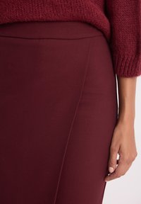 Bordowa, dopasowana spódnica z gładkiego materiału, z bocznymi szwami i pasem w talii. Miękka dziana bluzka częściowo widoczna na górze. Dłoń oparta na spódnicy.