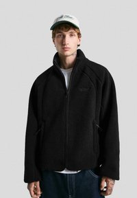 Jachetă din fleece neagră cu guler înalt, având un fermoar complet și buzunare laterale. Fabricată dintr-un material moale cu un finisaj texturat.