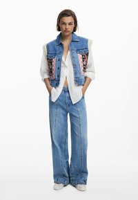 Denimweste mit Pelz-Schulterakzenten und floral gemusterten Seitenteilen, kombiniert mit hellblauen weiten Jeans und einem weißen Hemd mit Knopfleiste.