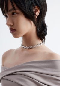 Silberne Ketten-Choker mit ineinandergreifenden Gliedern, getragen über einem glatten, drapierten, hellbeigen Stoff. Weiche Textur und minimalistische Designdetails.
