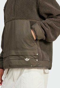 Fleece-Pullover in dunklem Olivgrün mit strukturiertem oberen Abschnitt und glattem unteren Teil, ausgestattet mit einem halben Reißverschluss, einer Seitentasche und Logo-Akzenten.