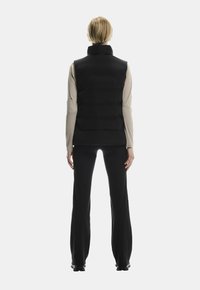 Sort puffer vest med høj krave, der har et kviltet design og letvægtsstof, båret over en lys langærmet top og sorte bukser.