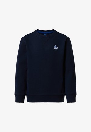 Jeune garçon portant un sweat-shirt et un pantalon bleu marine, des baskets blanches et une casquette orange North Sails, debout les mains dans les poches sur un fond uni.
