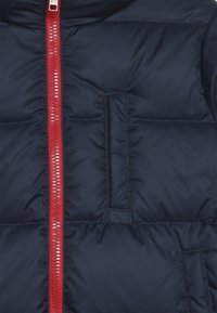 Nafukovací bunda v námořnické modři se středním červeným zipem a malou kapsou na levé straně. Hladký materiál s prošívanou strukturou.
