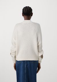 Maglione bianco a maglia con colletto alto e maniche larghe, indossato su una gonna in denim, con orlo a costine e vestibilità casual.