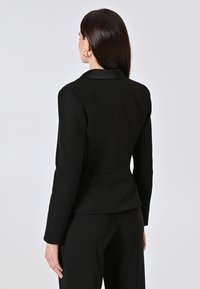 Blazer nero su misura con colletto a punte, maniche lunghe e vestibilità strutturata, caratterizzato da una texture liscia e un design minimalista.