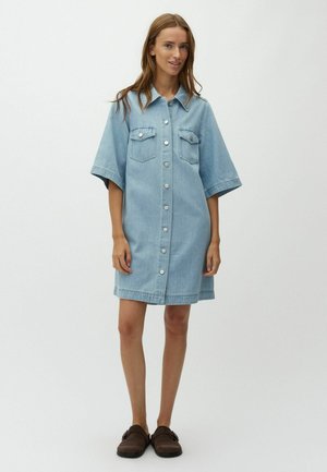 Frau trägt ein hellblaues Jeanskleid mit kurzen Ärmeln, zwei Brusttaschen und dunkelbraunen Slipperschuhe, steht vor einem einfarbigen Hintergrund.