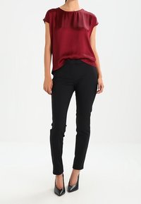 Top corto a maniche corte in satin bordeaux abbinato a pantaloni slim-fit neri e tacchi alti neri, in piedi contro uno sfondo chiaro.