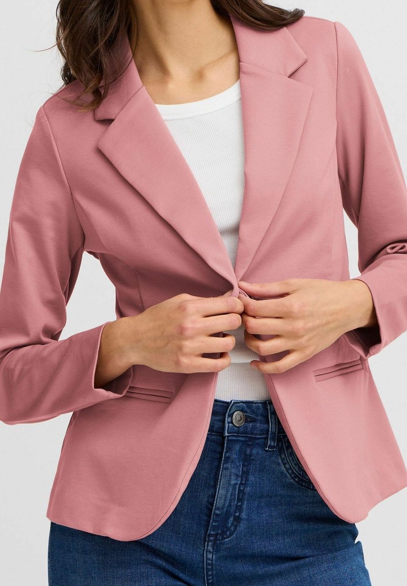 Femme portant un blazer rose sur une chemise blanche et un jean bleu, en train de boutonner le bouton avant du blazer.