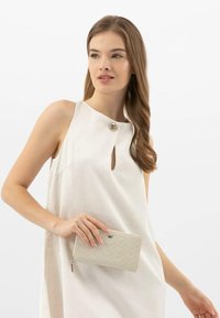 Světle béžové šaty bez rukávů s výstřihem ve tvaru klíče a stříbrným knoflíkem. Modelka drží texturovanou béžovou peněženku s vzorem v šeříkovém stylu.