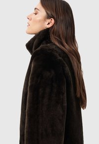 Casaco de pele sintética castanho escuro com gola alta e acabamento macio e texturizado. O casaco tem um corte solto, costuras visíveis e sem fechos à vista.