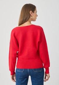 Pull rouge côtelé avec un large décolleté, de longues manches ballons et une taille cintrée, associé à un jean bleu.