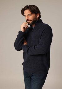 Pull-over en laine marine avec un motif texturé, col à demi-zip et poignets côtelés. Porté avec un jean foncé, il présente une coupe décontractée.