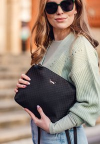 Černá texturovaná kabelka s geometrickým vzorem, s pozlaceným logem jako akcentem. Nosená s světle zeleným kardiganem a džíny.