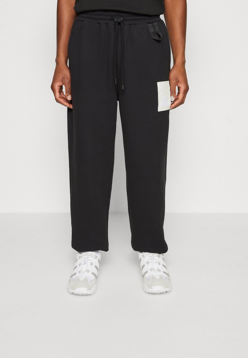 Pantalon de survêtement noir avec une taille élastique, un cordon de serrage réglable et une poche plaquée blanche sur la cuisse gauche. Texture de tissu lisse.