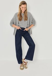 Jeans de mezclilla oscuros de pierna ancha, cárdigan de punto gris, top gris, zapatillas beige con acentos rojos. Pelo largo, manos en los bolsillos, atuendo casual.