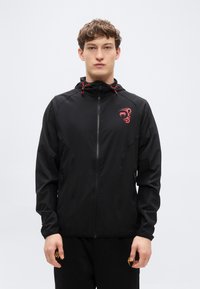 Μαύρο μπουφάν με φερμουάρ και κουκούλα, με κόκκινες κορδόνια και κόκκινο λογότυπο στο αριστερό στήθος. Ελαφρύ ύφασμα με λεία υφή.