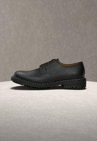Chaussure habillée pour homme en cuir noir à lacets, avec bout arrondi et semelle épaisse, posée sur une surface et un arrière-plan neutres.