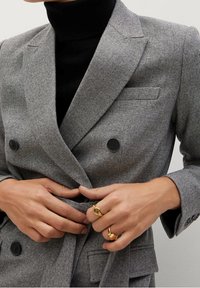 Des mains ajustent un blazer gris croisé porté sur un col roulé noir, avec des bagues en or aux doigts.