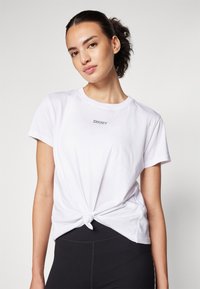 Balta kokvilnas T-kreklā ar mezglotu priekšpusi, uz krūtīm ir neliels pelēks DKNY logo. Saskaņots ar melniem legingiem.