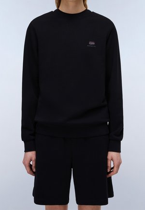 Person iført en sort langærmet sweatshirt og matchende sorte shorts med et lille logo på den øverste venstre side af sweatshirten.