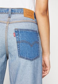 Světle modré džíny z denimu s hnědým koženým logem, oranžovým šitím a výrazným designem zadní kapsy s modrým vsazením.
