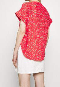 Czerwona koszula z krótkim rękawem w białe kropki, z asymetrycznym dołem i luźnym krojem. Wykonana z lekkiego materiału o miękkiej teksturze.