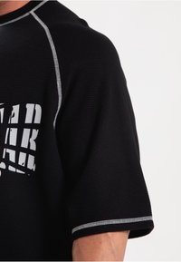 Zwart kortarm T-shirt van gestructureerde stof, met witte bedrukte tekst en grijze stiksels langs de halslijn en mouwen.