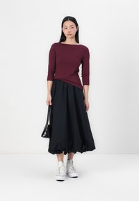 Burgundy, figurnära topp med trekvartslånga ärmar, kombinerad med en svart, voluminös kjol. Modellen bär vita sneakers och håller en liten svart väska.
