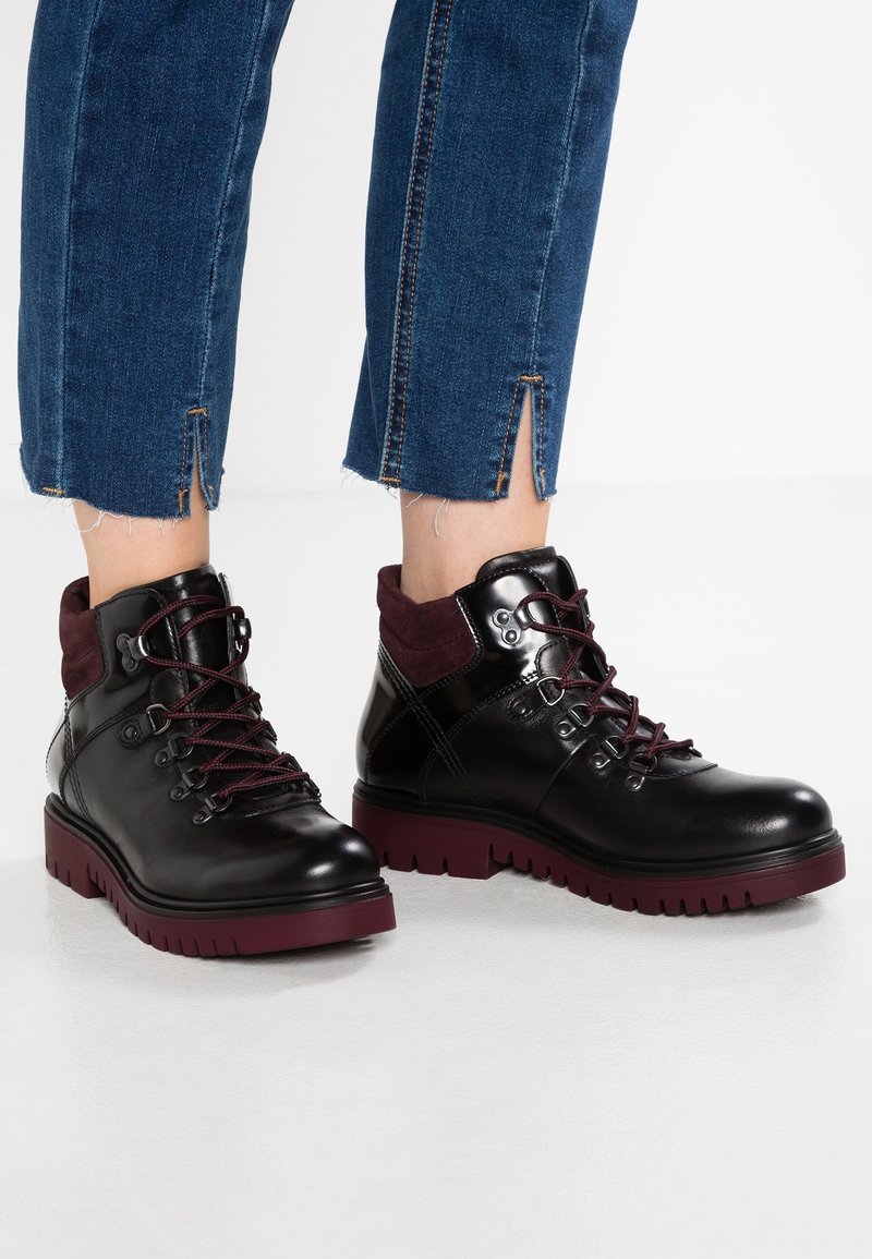 Bottes cheville en cuir noir avec semelles et lacets bordeaux, portées avec un jean bleu à fentes à la cheville.