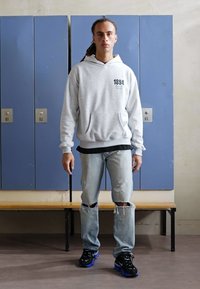 Hoodie gris avec poches avant, couche de base noire, jeans déchirés bleu clair, et baskets noires avec accents bleus, se tenant devant des casiers.