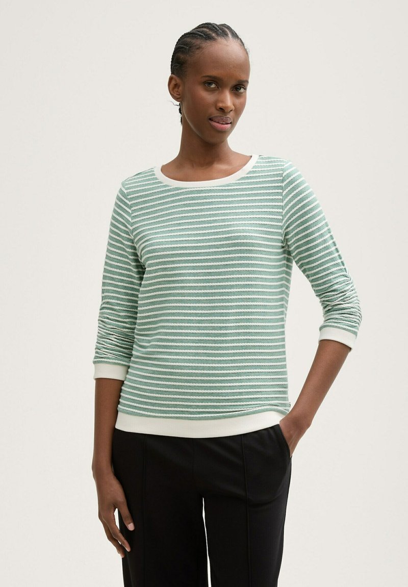 Hellgrüner gestreifter Pullover mit weißen Akzenten, rundem Ausschnitt und Dreiviertelärmeln, kombiniert mit schwarzen Hosen. Weiche Stofftextur.