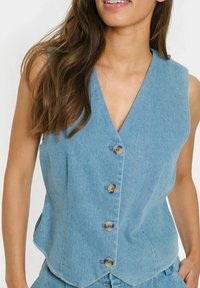 Jeans brezrokavnik v svetlo modri barvi z V-izrezom, petimi gumbi spredaj in poševnim robom. Gladka tekstura z rahlim leskom.