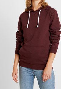 Donna che indossa un hoodie bordeaux con lacci bianchi e una tasca a marsupio, abbinato a jeans di denim azzurro chiaro, su uno sfondo semplice.