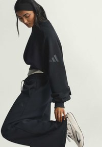 Žena u crnoj Adidas majici dugih rukava i hlačama s bijelim tenisicama, podešava nogavicu dok gleda prema dolje na jednobojnoj pozadini.