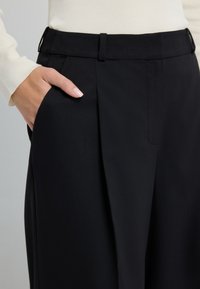 Zwarte geplooide broek van gestructureerde stof, met een op maat gemaakte tailleband en een zak aan de zijkant, afgewerkt met een gladde afwerking.