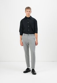 Jonge man die staat in een zwarte overhemd met opgerolde mouwen, lichtgrijze broek, witte sokken en zwarte nette schoenen tegen een witte achtergrond.
