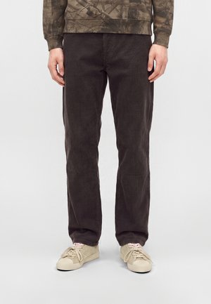 Man draagt donkerbruine corduroy broek, beige sneakers met roze veters en een groene trui met camouflagepatroon tegen een effen achtergrond.