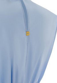 Vêtement en tissu bleu clair avec un design simple, comportant une couture subtile et une petite étiquette dorée près de l'encolure. Texture lisse.