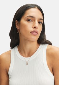 Guldtonade hoopörhängen med en rektangulär berlock, tillsammans med ett fint kedjehalsband med en liten berlock, bärs över en vit ribbad topp.