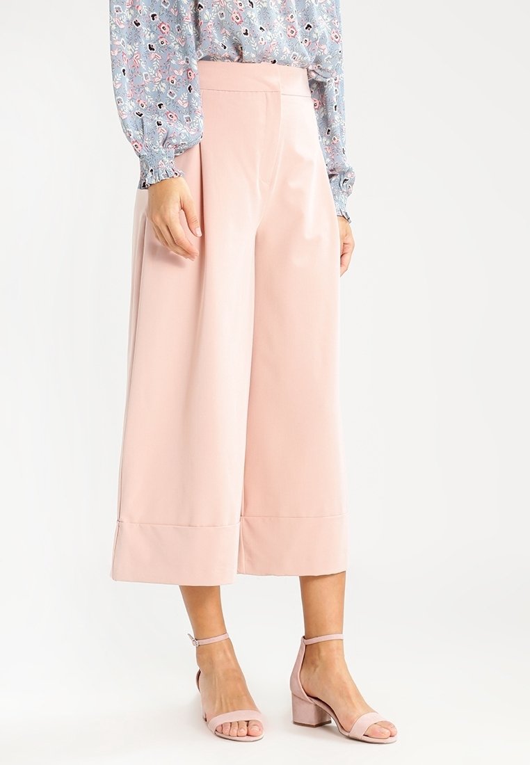 Femme portant un pantalon ample court de couleur rose pâle, un chemisier à manches longues à motifs floraux, et des sandales à talons carrés assorties rose.
