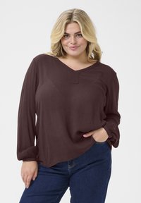 Brown long-sleeve blouse with a V-neck and a button detail at the neckline; light, textured fabric; paired with dark blue jeans.