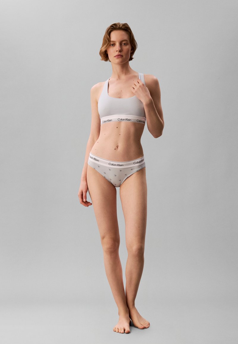 Γυναίκα με γκρι ανοιχτό Calvin Klein αθλητικό σουτιέν και αντίστοιχο εσώρουχο, στέκεται ξυπόλητη μπροστά σε απλό γκρι φόντο.