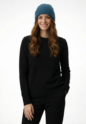 Young woman with long wavy hair smiling, wearing a blue knit beanie, black sweater, and black pants against a plain white background.